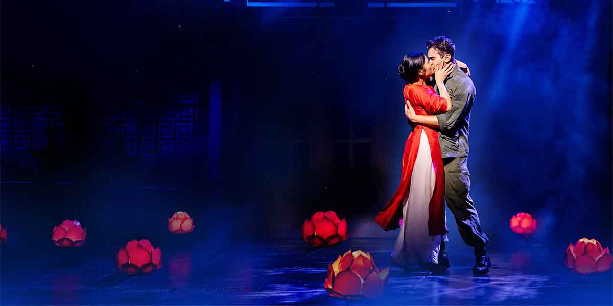 A couple embraces and kisses on stage, surrounded by glowing red lotus flowers and blue lighting.