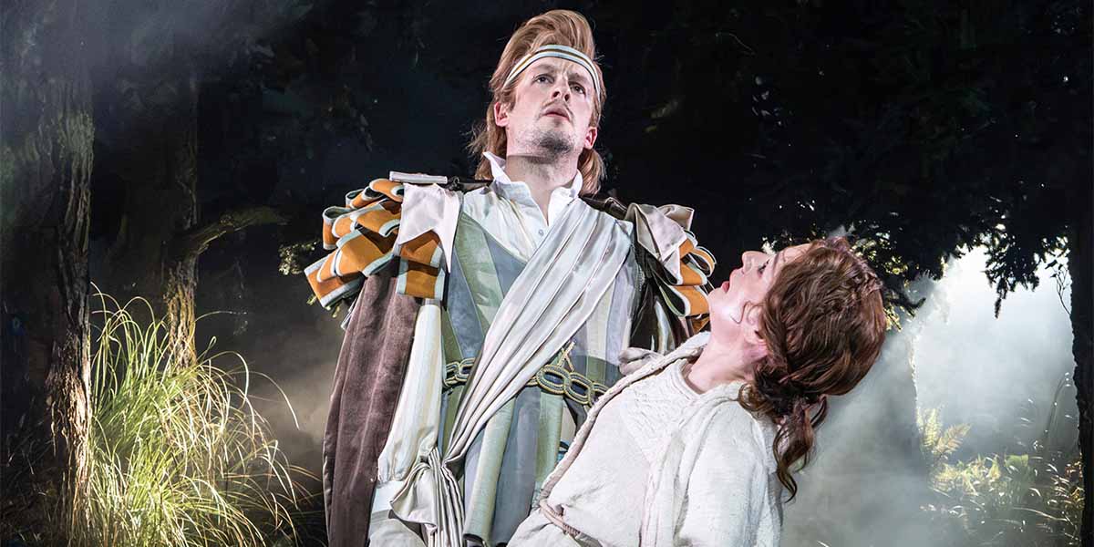 Matthew Seadon-Young and Laura Pitt-Pulford pose together in a forest setting, surrounded by lush greenery.