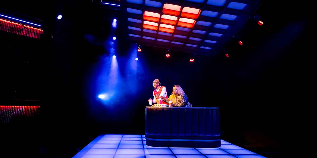 Michael Rishawn and Marisha Wallace perform at a table under dramatic blue and red stage lighting.