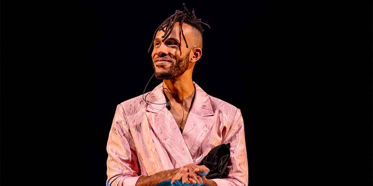 Michael Rishawn, wearing a pink patterned jacket, smiles while holding a black item on stage.