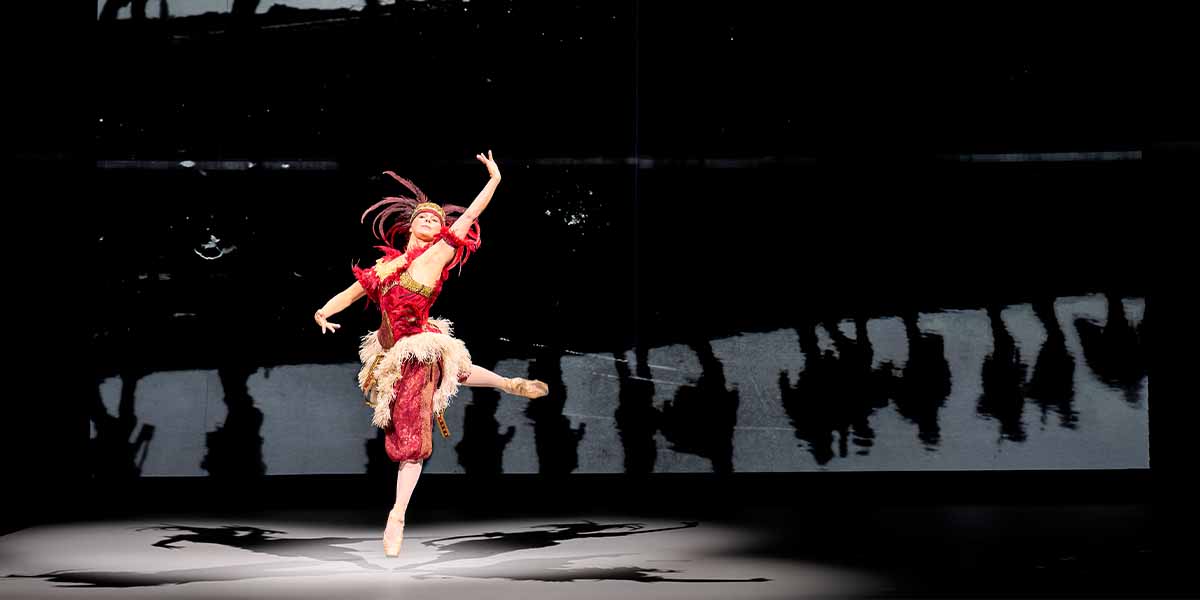 Natalia Osipova performs in a vibrant red costume, leaping gracefully against a dark background.