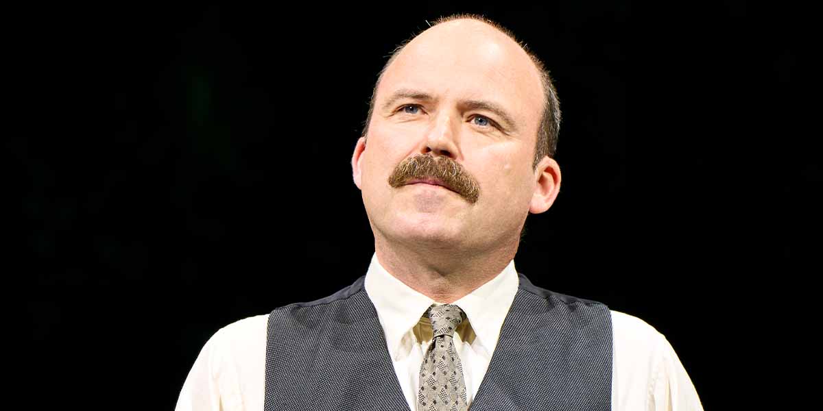 Rory Kinnear, wearing a white shirt and dark vest, gazes thoughtfully against a black background.