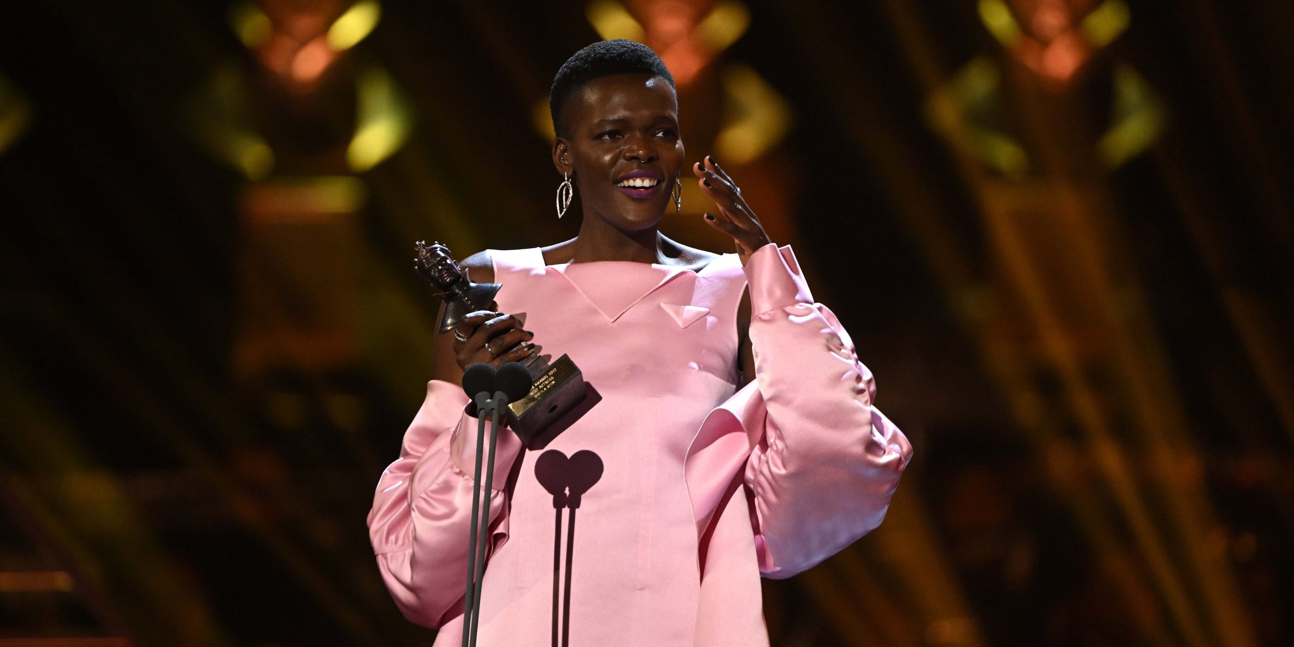 Sheila Atim poses confidently, wearing a black top, against a neutral background with soft lighting.