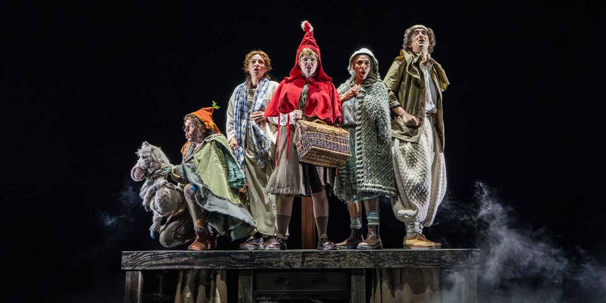Five performers in costumes stand on a wooden platform, with a foggy background, portraying characters from "Into The Woods.