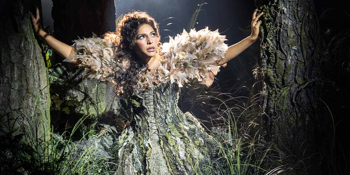 Simbi Akande as Cinderella, wearing a blue gown, stands in a forest setting, surrounded by trees.