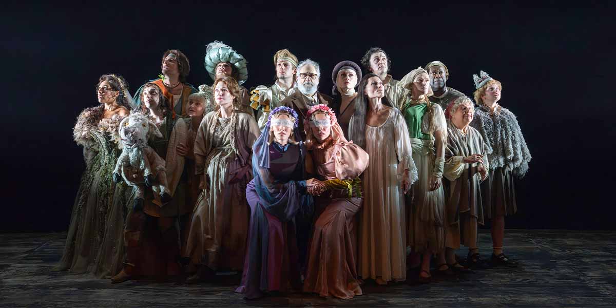 The cast of "Into The Woods" poses together in varied costumes against a dark background.