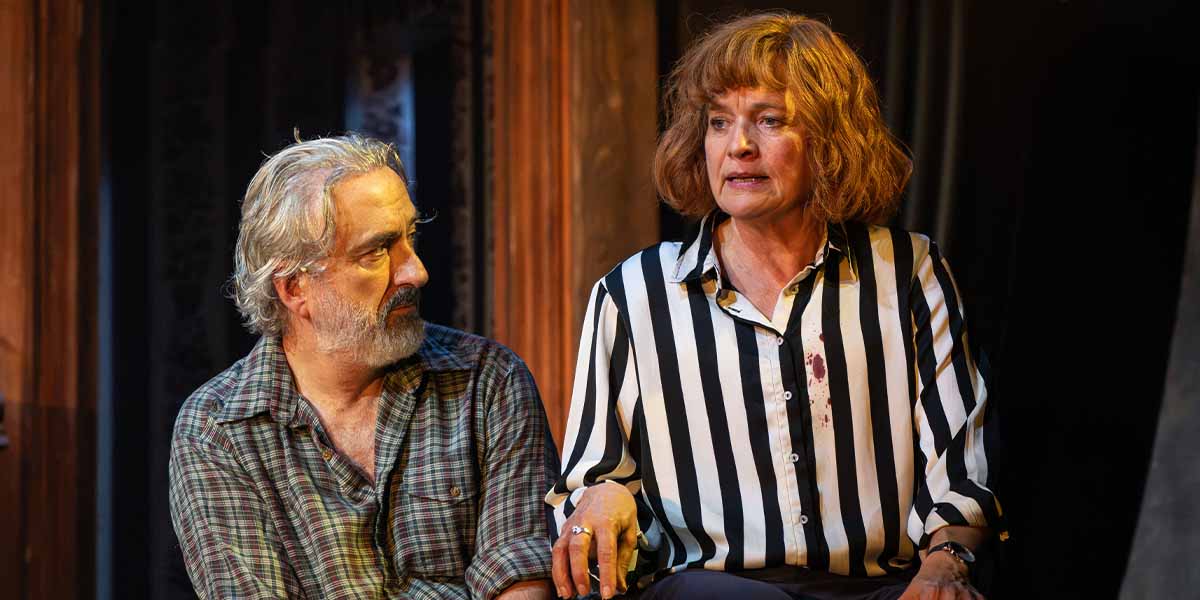 Declan Conlon and Kerry Fox sit together, engaged in conversation, with warm lighting and dark background.