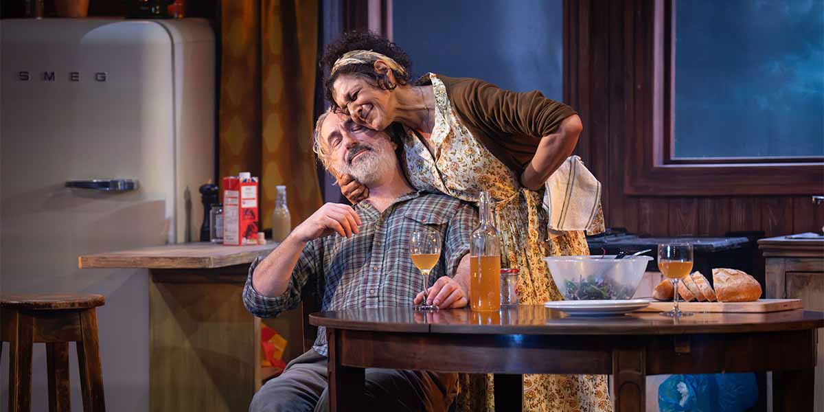 Declan Conlon and Meera Syal share an intimate moment at a kitchen table, surrounded by food and drinks.