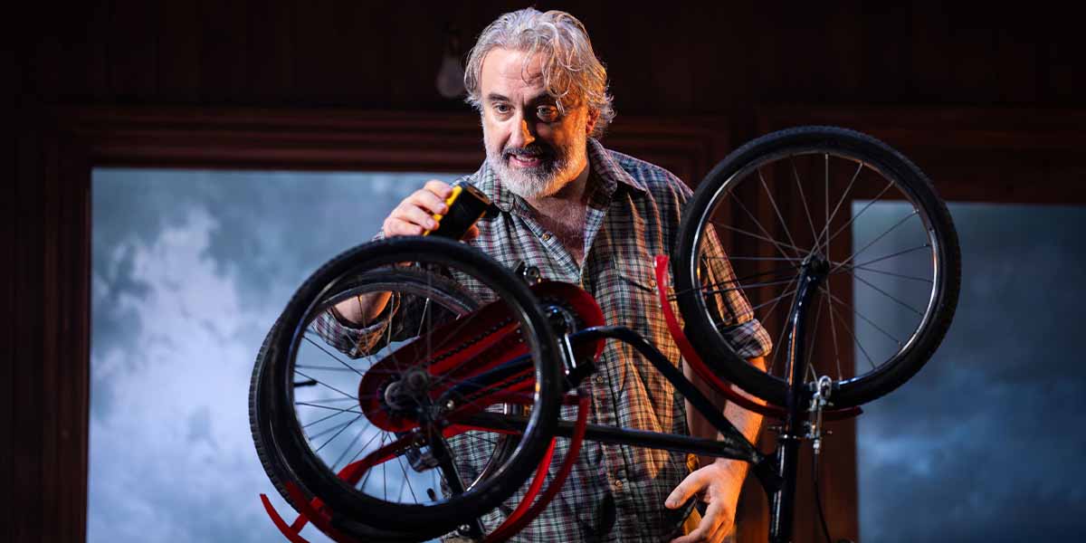 A man with grey hair holds a can while inspecting a bicycle in a dimly lit room.