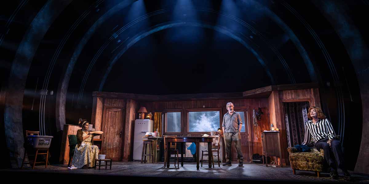 Three actors perform on stage in a wooden room, with dramatic lighting and a cloudy backdrop.