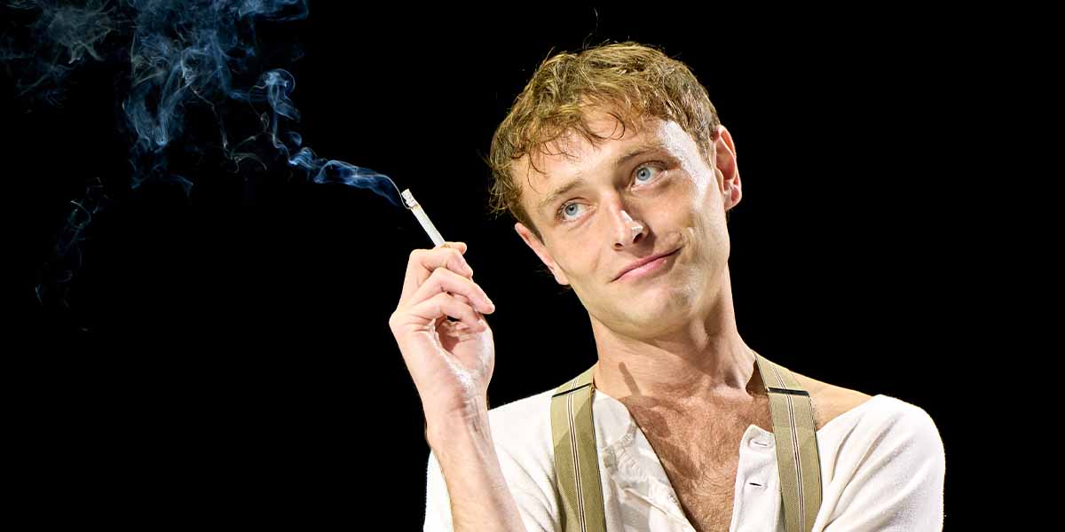 Thomas Flynn holds a cigarette, exuding a contemplative expression against a black background, with smoke curling upwards.