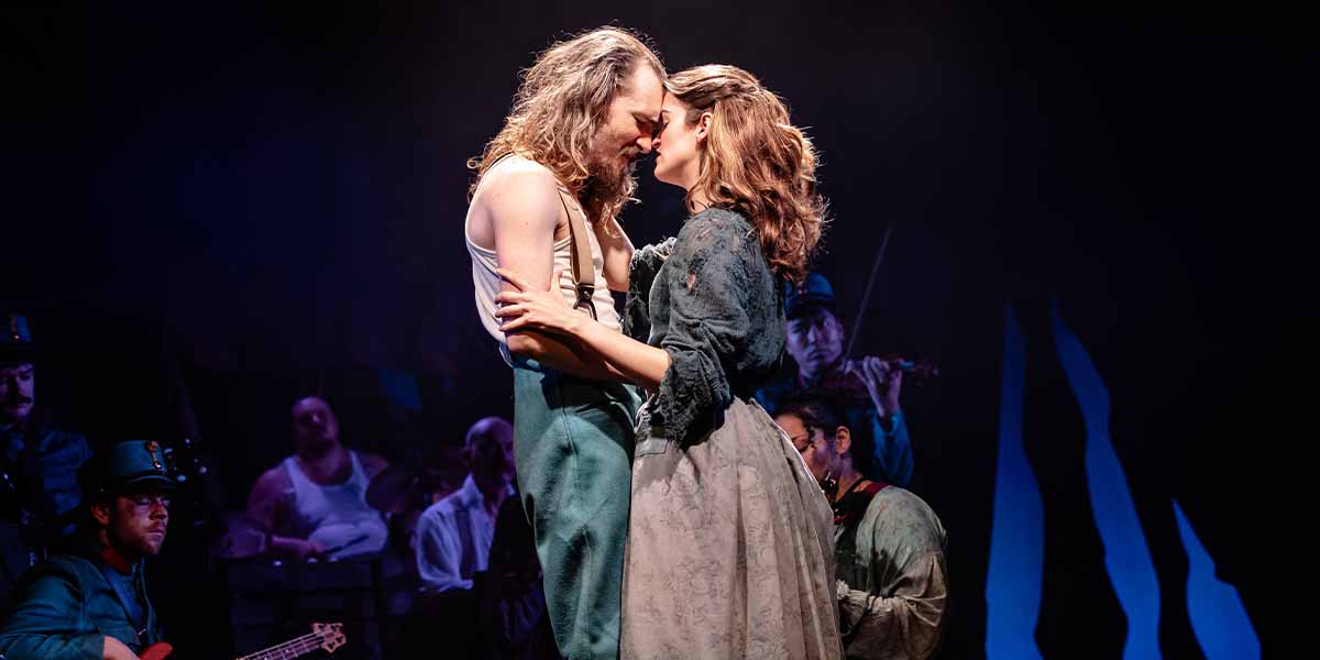 Franz and Marie embrace closely, their foreheads touching, with musicians visible in the dark background.