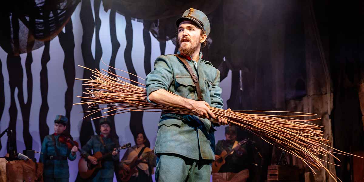A soldier in a blue uniform holds a bundle of twigs, with musicians visible in the background.