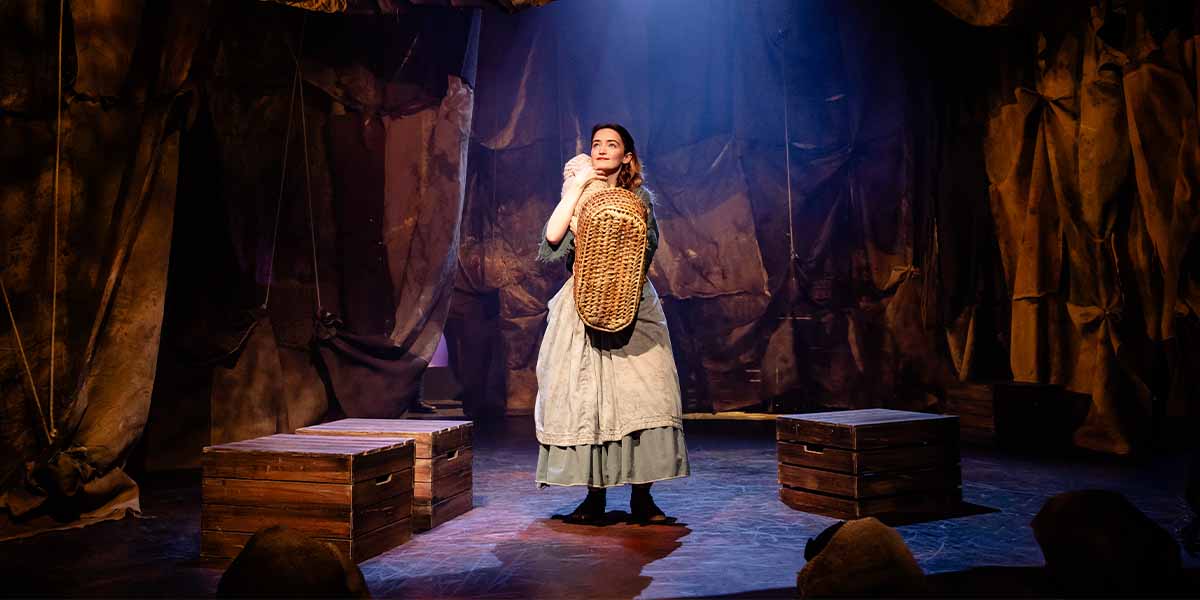 A woman in period costume holds a basket, standing on stage with wooden crates and dramatic lighting.