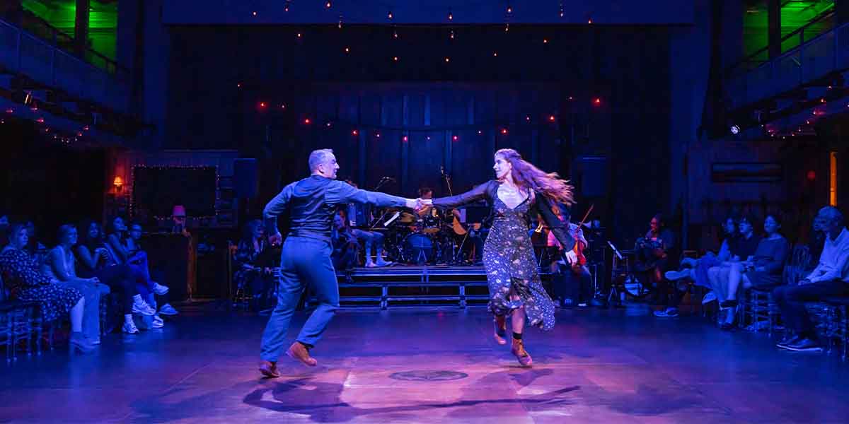 A man and woman dance together on stage, surrounded by an audience, with vibrant lighting and instruments.