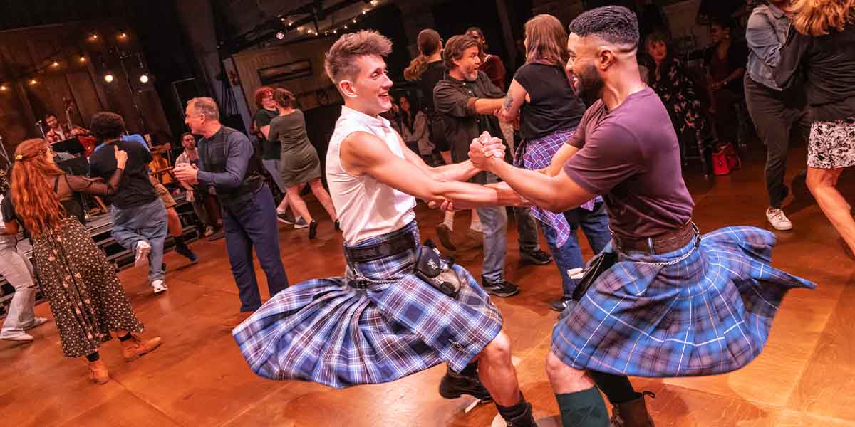 Two male dancers in kilts engage in a lively ceilidh dance, surrounded by a vibrant crowd.