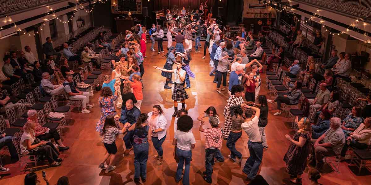 The cast of CEILIDH poses together, wearing vibrant costumes, smiling against a colourful backdrop.