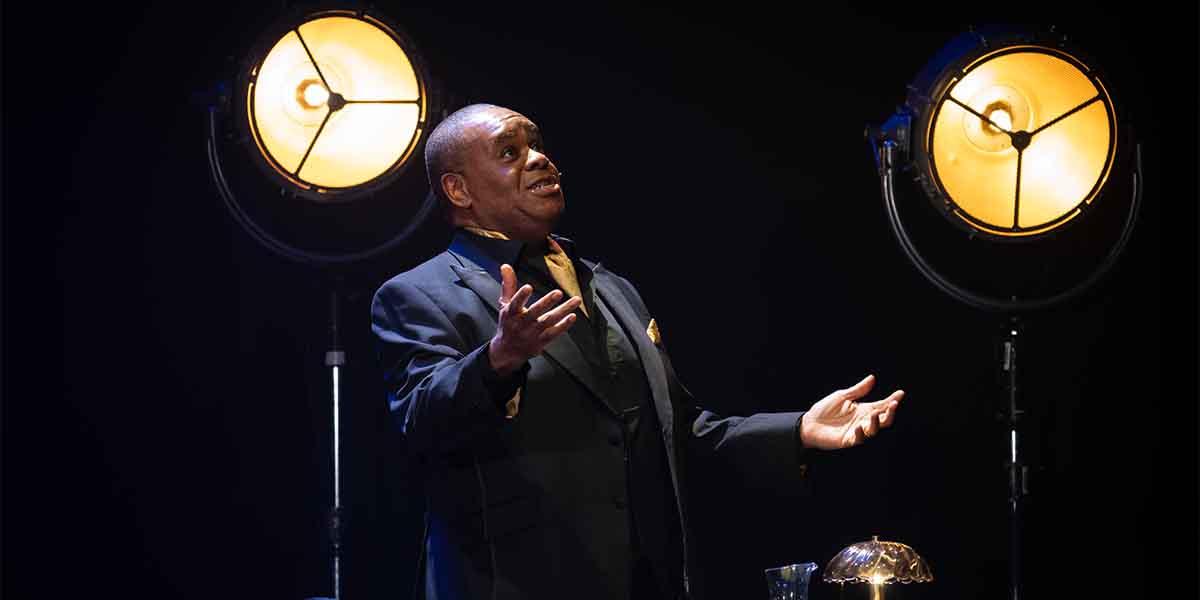 A man in a black suit gestures expressively, illuminated by warm stage lights, against a dark background.