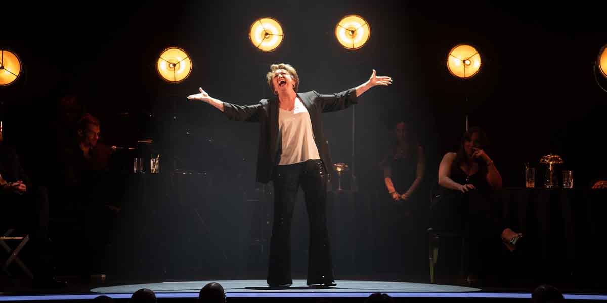 A performer in a black blazer and sequined trousers gestures passionately on stage, illuminated by spotlights.