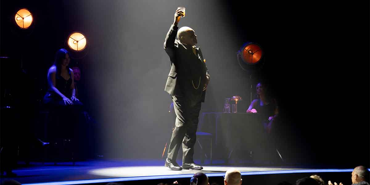 A performer in a black suit raises a glass on stage, with seated audience members and warm lighting.
