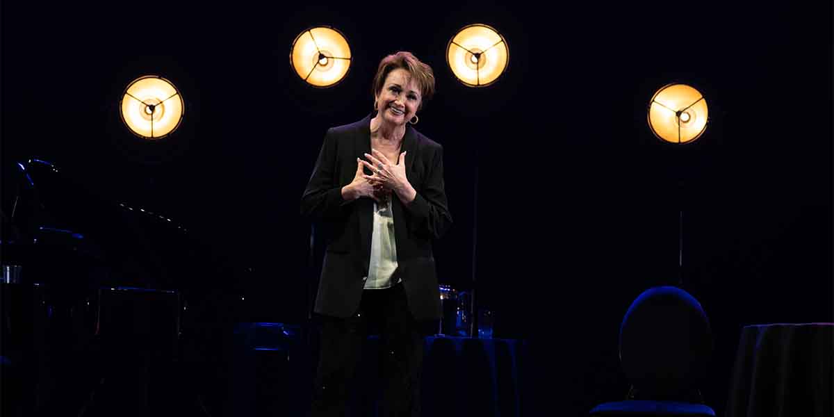 A woman in a black blazer smiles warmly, gesturing with her hands, under soft stage lighting.