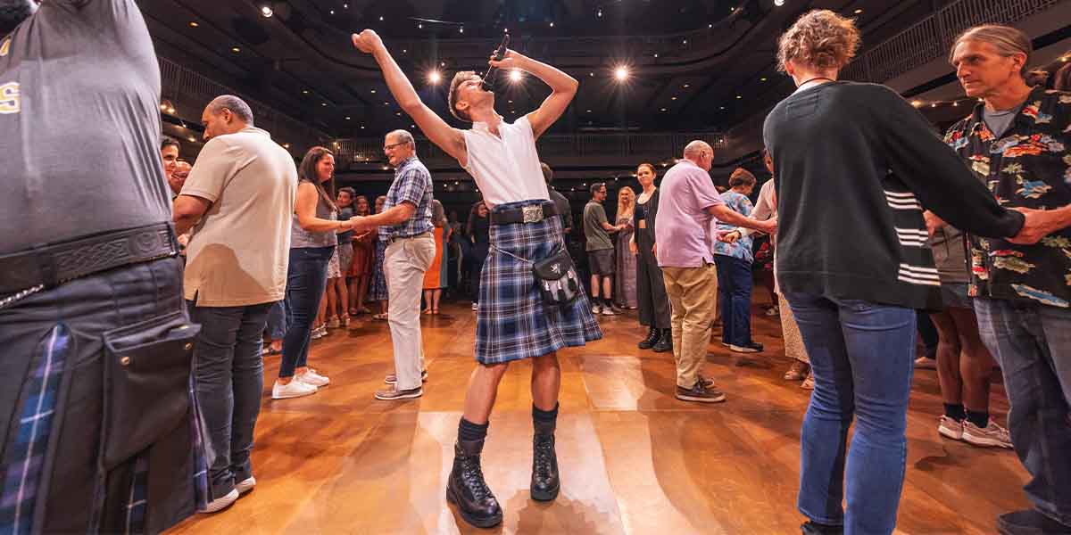 Scott Gilmour, in a kilt, performs energetically while others dance and interact in a lively setting.