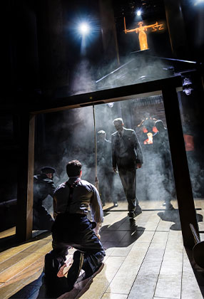A dramatic stage scene with smoke, featuring a man in a suit and two kneeling figures.