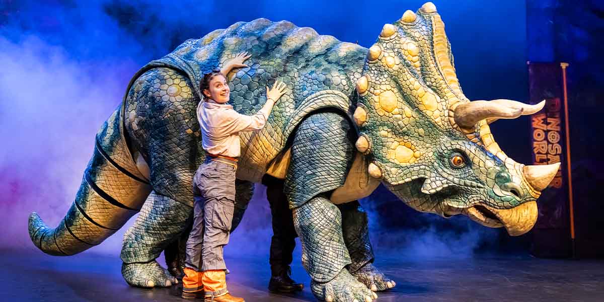 Max Humphries interacts with a large dinosaur puppet, surrounded by a vibrant stage set with greenery.