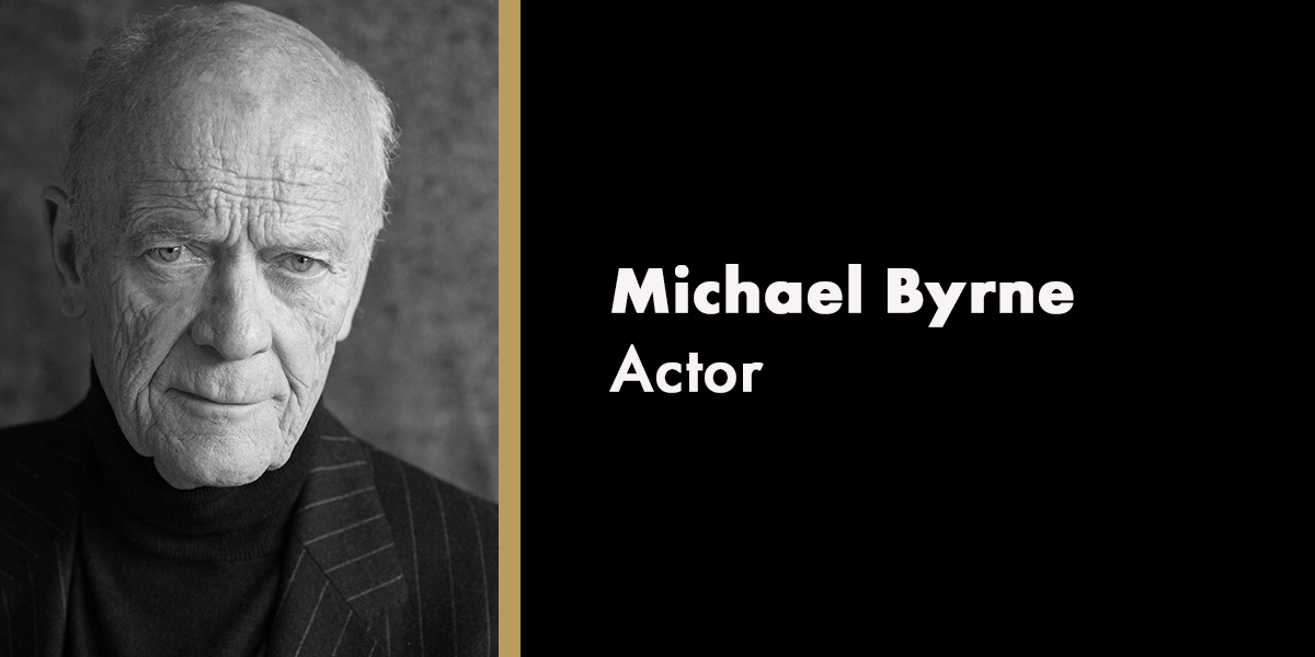 A portrait of Michael Byrne, smiling, wearing a dark suit against a neutral background.