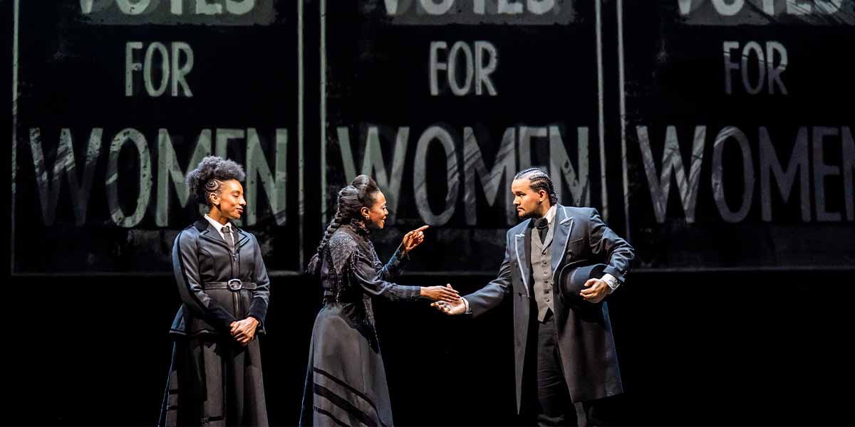 Three actors on stage, dressed in period costumes, engage in a handshake against a backdrop promoting women's suffrage.