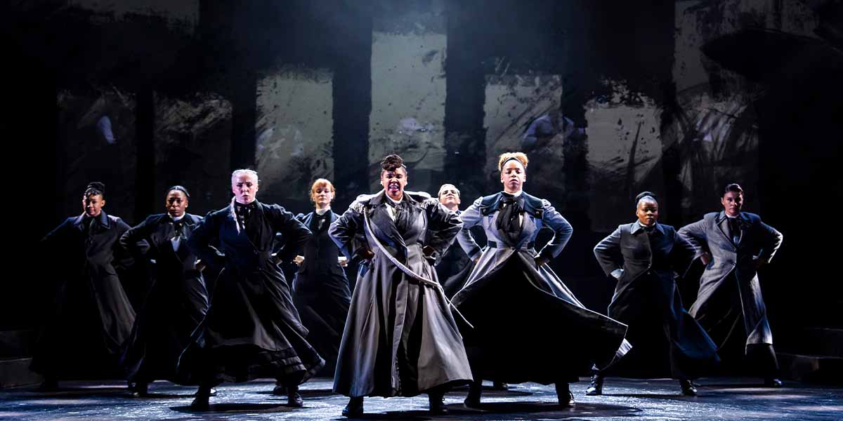 Nine performers in dark costumes pose dramatically on stage, with a textured backdrop enhancing the scene.