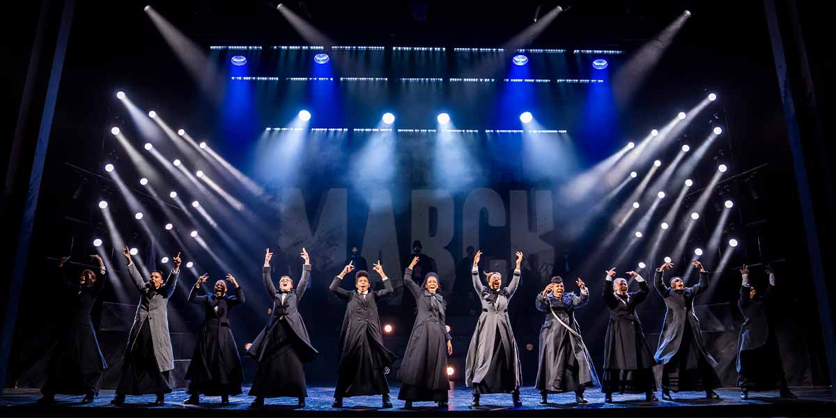 A group of performers in black coats pose dramatically on stage, illuminated by bright stage lights.