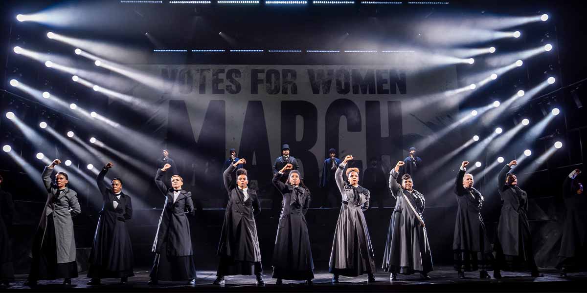 Eight performers in black costumes raise their fists in solidarity against a backdrop reading "Notes for Women March.
