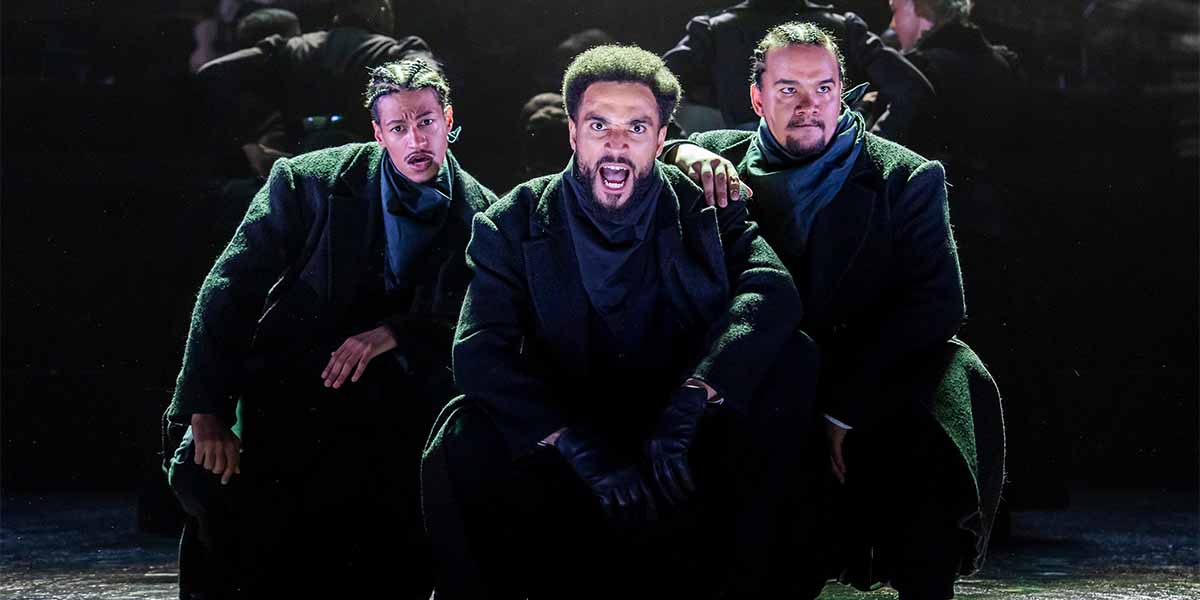 Three men in dark coats and scarves pose dramatically on stage, with intense expressions and gestures.