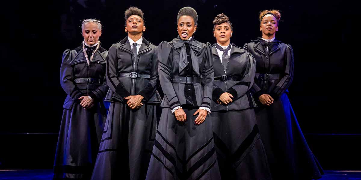 Five women in black Victorian-style dresses stand in a row, with dramatic expressions against a dark background.