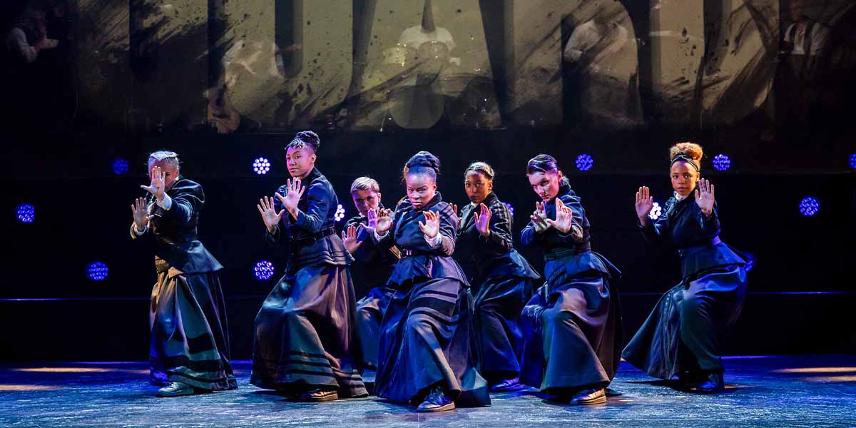 Seven dancers in black costumes pose dramatically on stage, with blue lights illuminating the background.