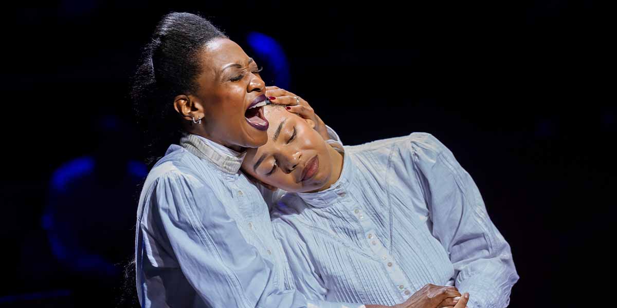 Two women in white blouses embrace, one singing passionately while the other rests her head gently.