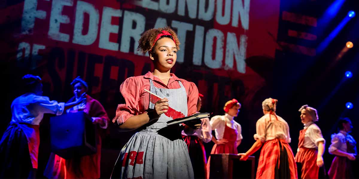 A young woman in a red blouse and apron holds a notebook, with performers in the background.