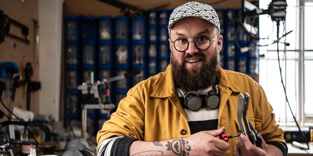 Max Humphries smiles while holding tools in a workshop, wearing a yellow jacket and patterned cap.