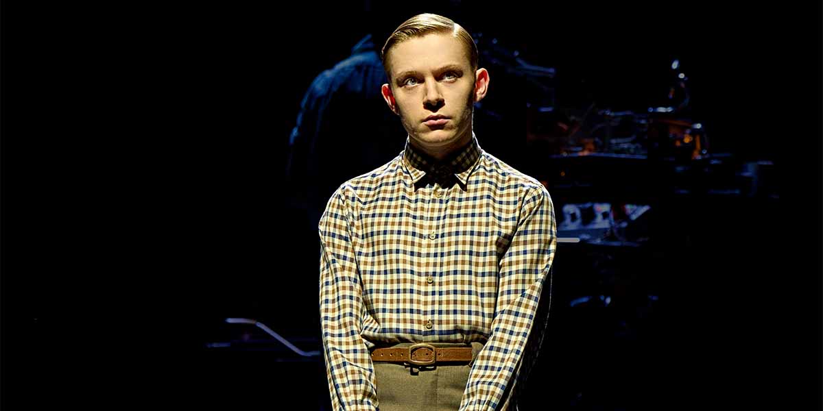 Paddy Bever stands with a serious expression, wearing a checked shirt and trousers against a dark background.