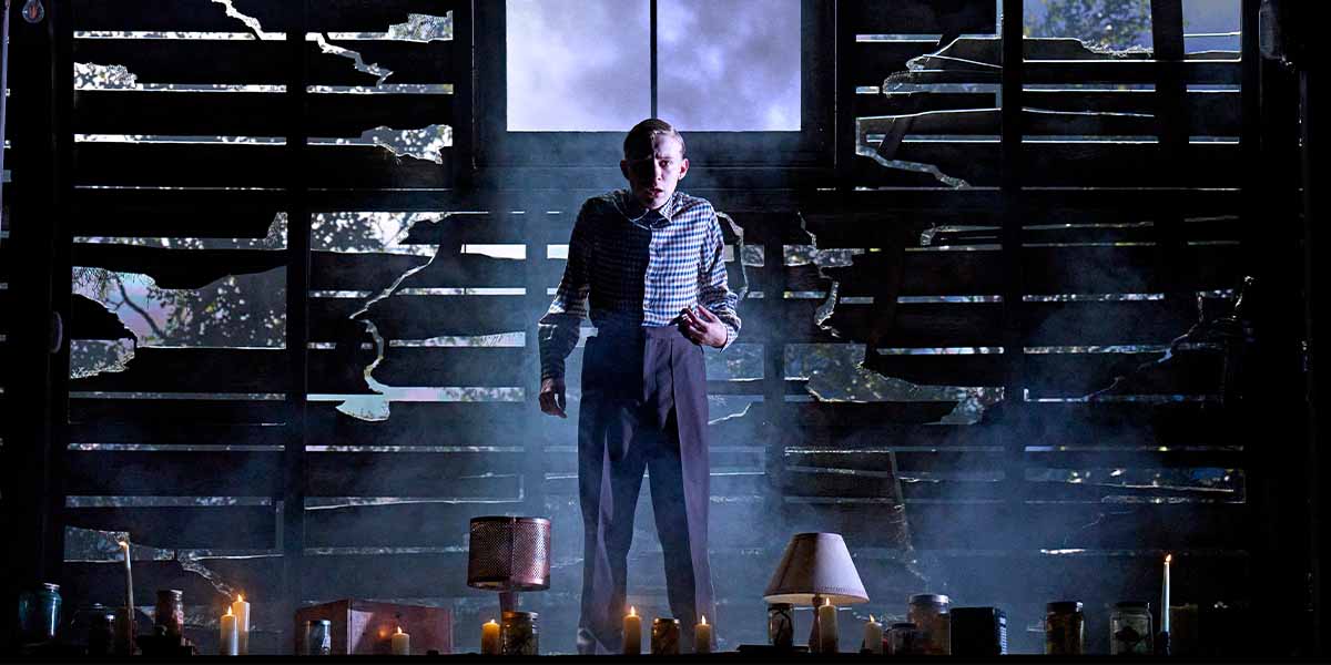 Paddy Bever stands in a dimly lit, rustic space, surrounded by candles and broken wooden panels.