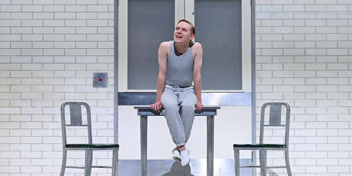 Paddy Bever sits on a table, smiling, with two empty chairs and a white tiled wall behind.