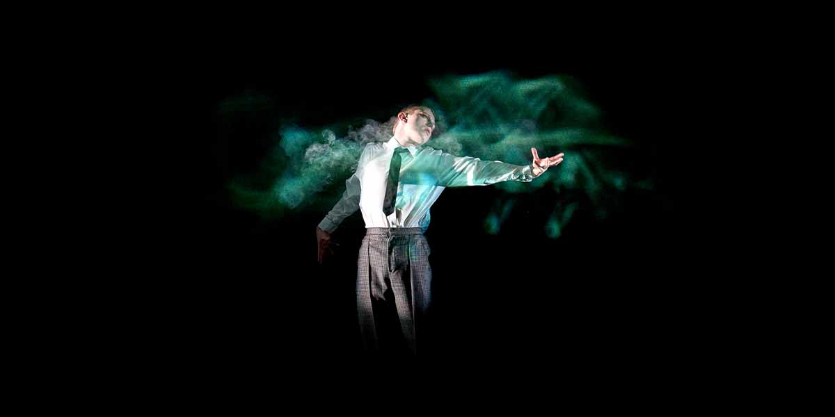 Paddy Bever poses dramatically in a white shirt and tie, surrounded by swirling green smoke against a dark background.