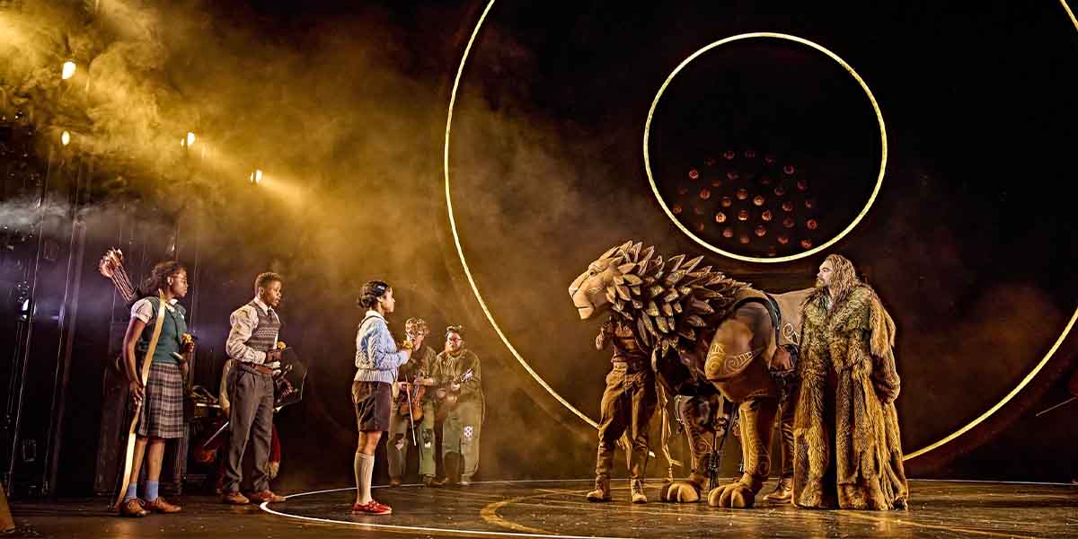 A lion puppet stands centre stage, surrounded by actors in costumes, with dramatic lighting and circular backdrop.