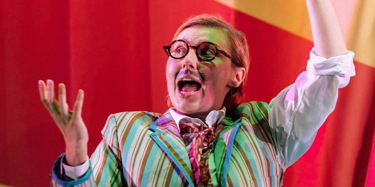 Bronté Barbé, wearing a striped suit, passionately gestures while performing on stage with a vibrant backdrop.