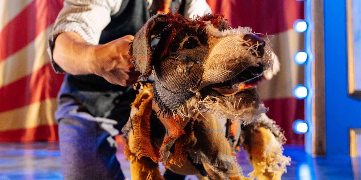 Calum Donald-Bruce performs with a textured puppet dog against a vibrant, striped backdrop at the theatre.