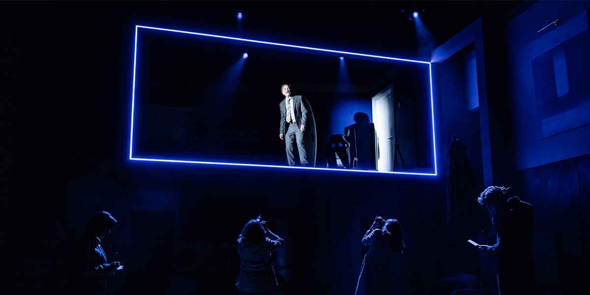 Donald Sage Mackay as Charles stands in a spotlight, surrounded by darkened figures taking photos.