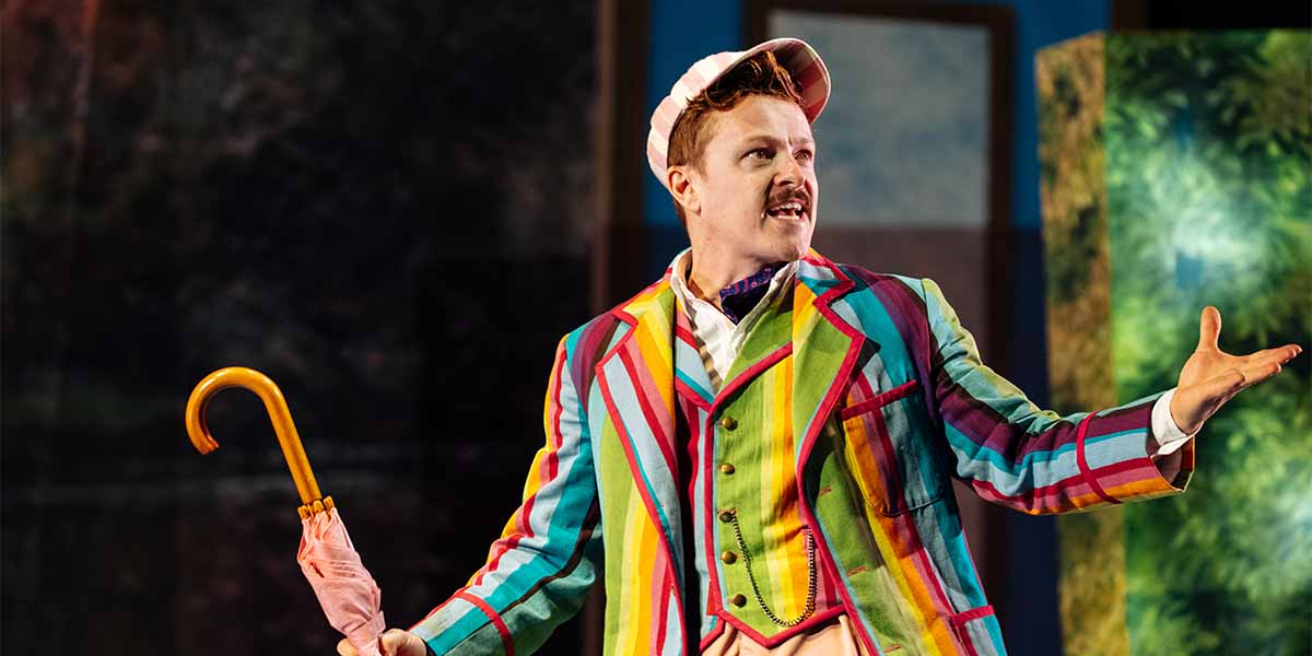 Jonny Weldon as Jerome, wearing a multicoloured striped suit, gestures animatedly with a pink umbrella.