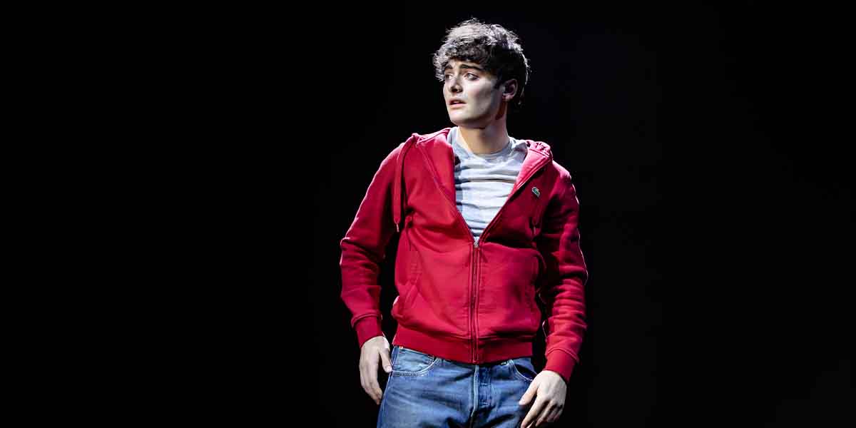 Noah Schnapp as Dylan, wearing a red hoodie and jeans, stands on stage against a dark background.