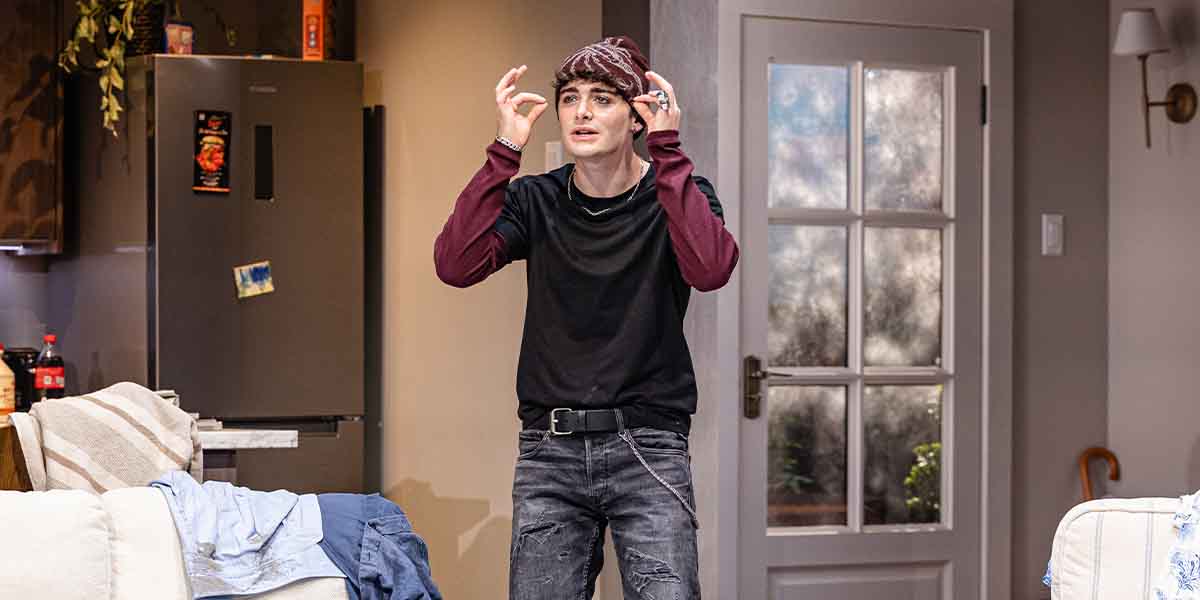 Noah Schnapp, as Todd, gestures expressively in a domestic setting with a fridge and sofa visible.